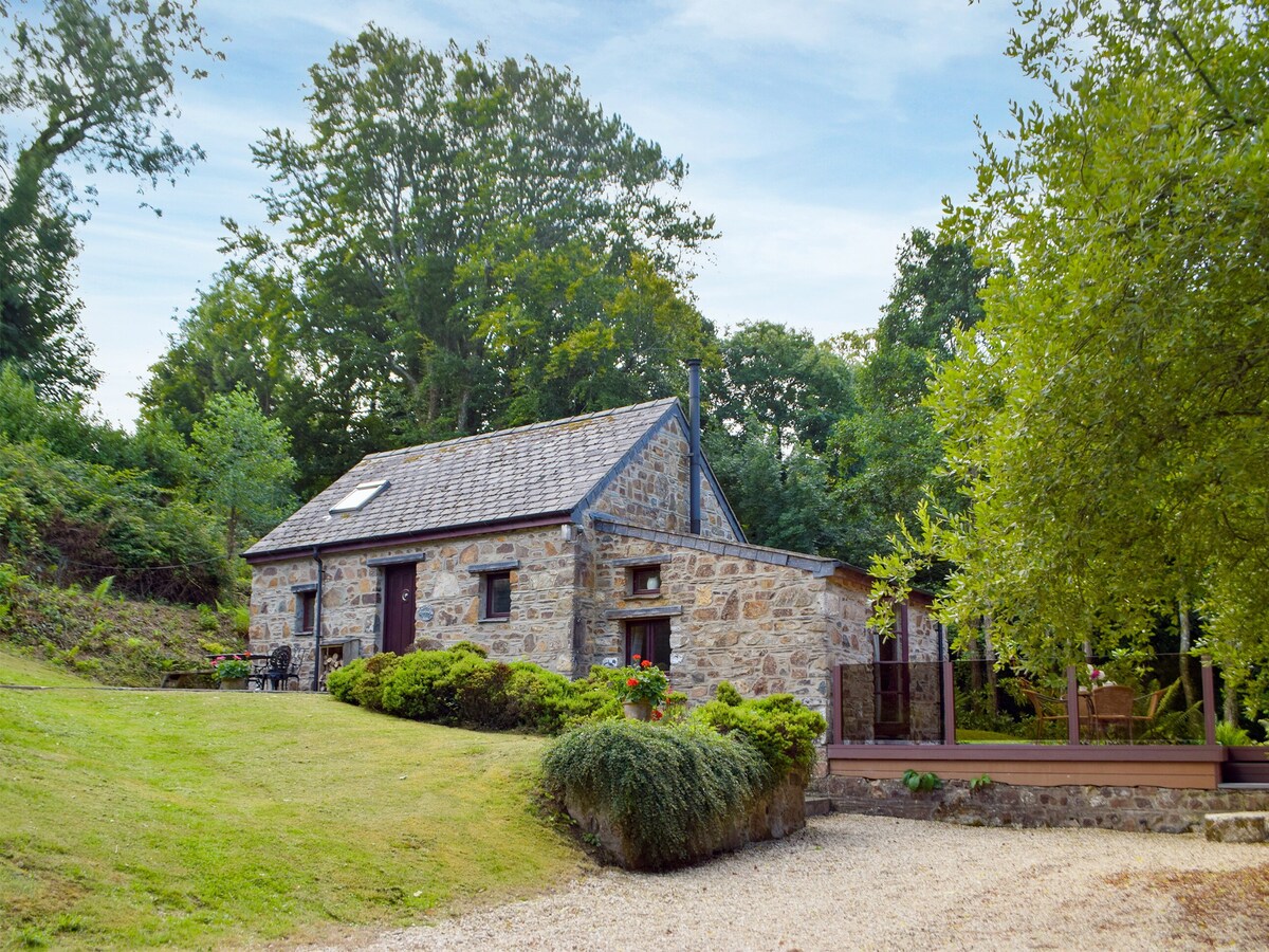 A charming stone cottage is nestled amidst greenery, with a sloped lawn leading to the entrance. Large trees and shrubs surround the exterior, complementing the cottage's rustic character. The roof features a skylight, and a small seating area is visible in front of the cottage.