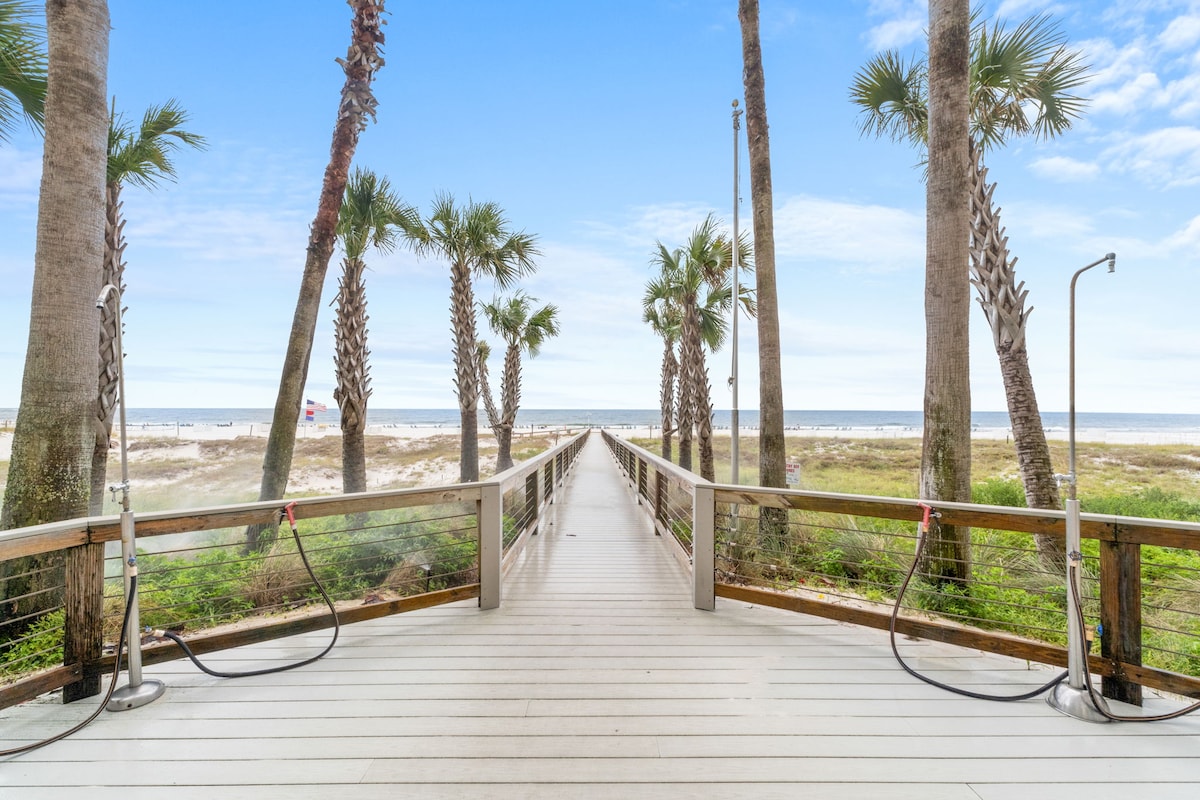 A wooden boardwalk leads directly to the sandy beach, flanked by palm trees that provide a tropical ambiance. The clear blue sky and gentle waves create a peaceful coastal setting, inviting guests to step outside and enjoy the beach access.
