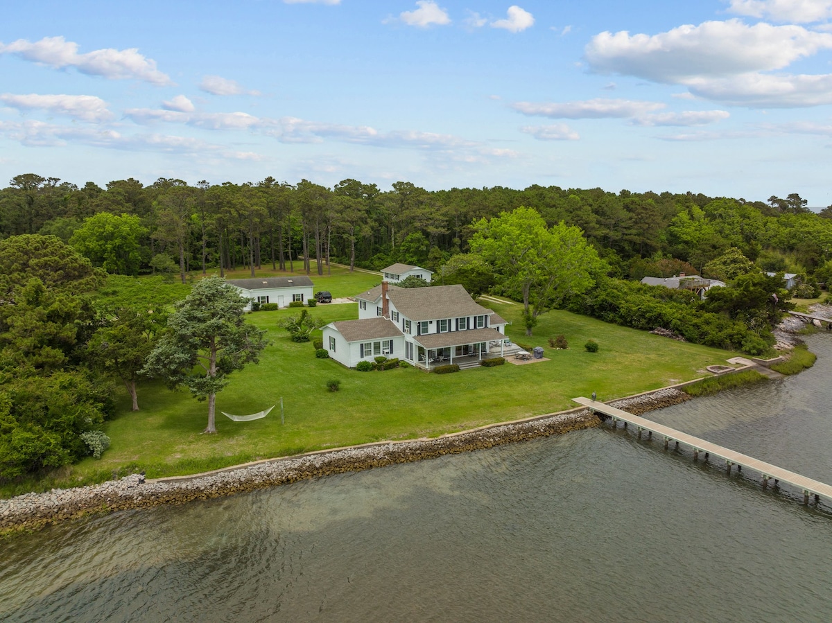The exterior view of Harold's House showcases a charming two-story home nestled among lush trees and expansive greenery. A private dock extends into the water, with a hammock placed in the yard offering a serene spot for relaxation. Scenic water views surround the property.