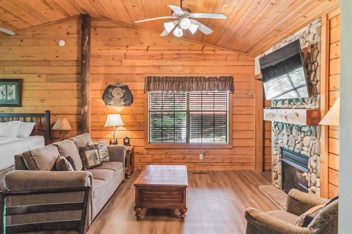 A warm living area features wooden paneling and a vaulted ceiling with a ceiling fan. A comfortable sofa with decorative pillows is positioned near a coffee table. A stone fireplace anchors one side, while a mounted television adorns the wall above.