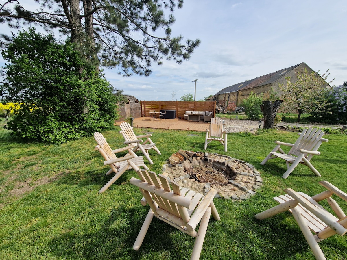 A circular stone fire pit is surrounded by several light-colored wooden chairs on a grassy area. In the background, a wooden deck can be seen, equipped with seating and framed by green foliage and blooming trees.