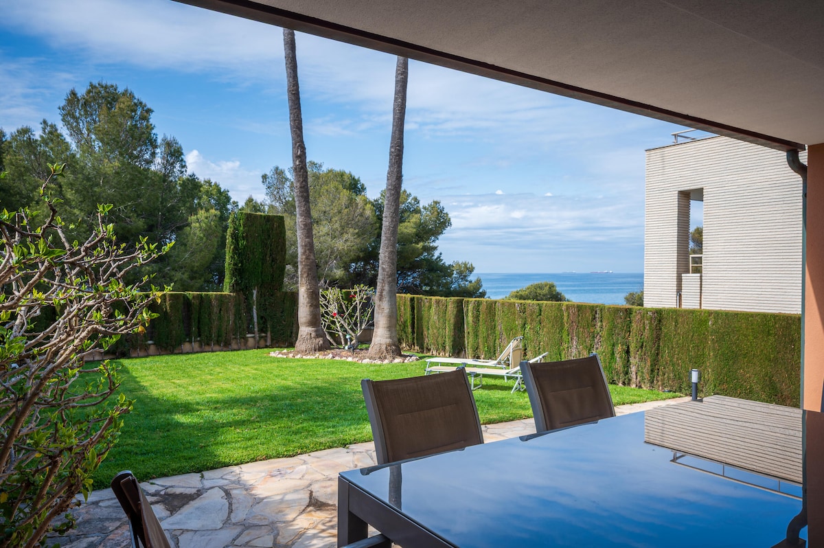 A spacious outdoor terrace is presented, featuring a dining table paired with modern chairs. Lush greenery surrounds the area, with neatly trimmed hedges and palm trees adding a tropical feel. The serene view of the ocean is visible in the background, enhanced by a clear blue sky.