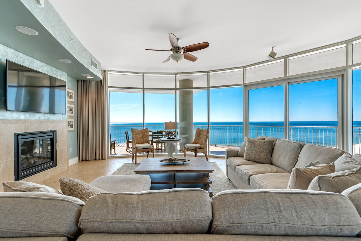 An open and airy living space features a cozy sectional sofa facing a flat-screen TV and a fireplace. Floor-to-ceiling windows frame breathtaking Gulf views. A circular dining table is positioned nearby, underscoring the inviting atmosphere of this stylish room.