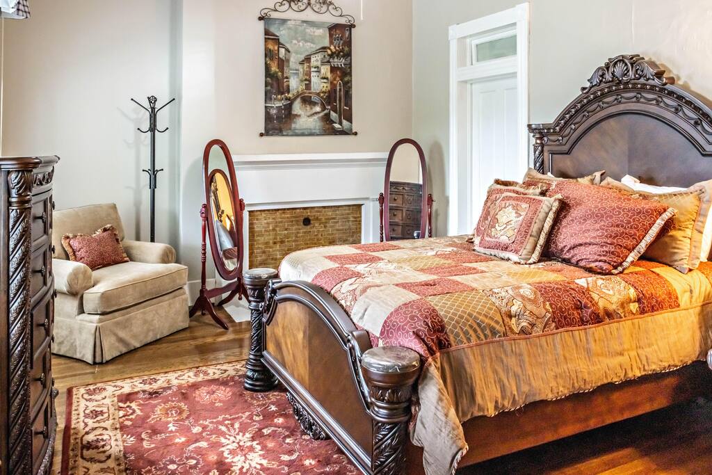 The room features a spacious bed adorned with rich, patterned bedding in warm tones. A cozy chair is positioned nearby, alongside a decorative area rug and mirrors enhancing the elegance of the space. Natural light filters in through the windows, creating a serene atmosphere.