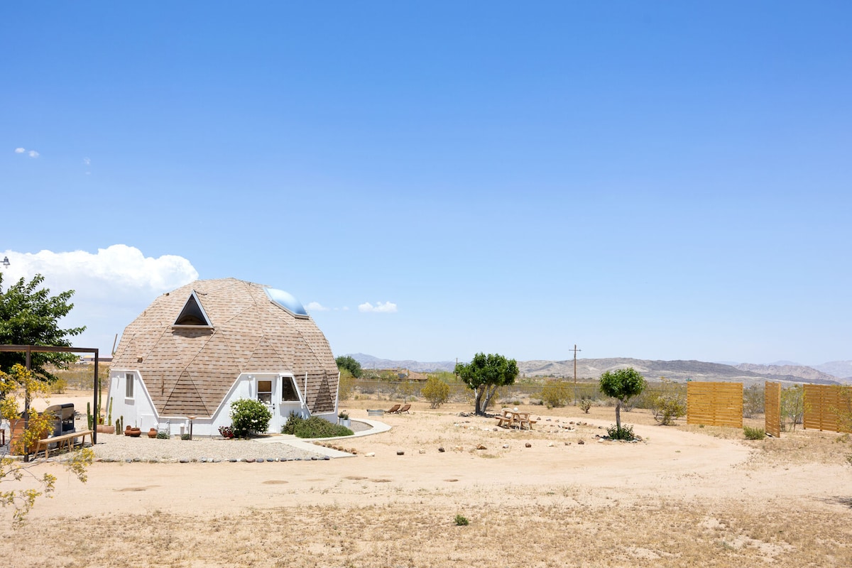 A geodesic dome home is set against a clear blue sky, surrounded by sprawling desert landscape. The structure features distinctive triangular windows and is framed by low desert vegetation. Nearby, a circular area is visible, indicating outdoor seating or lounging space.