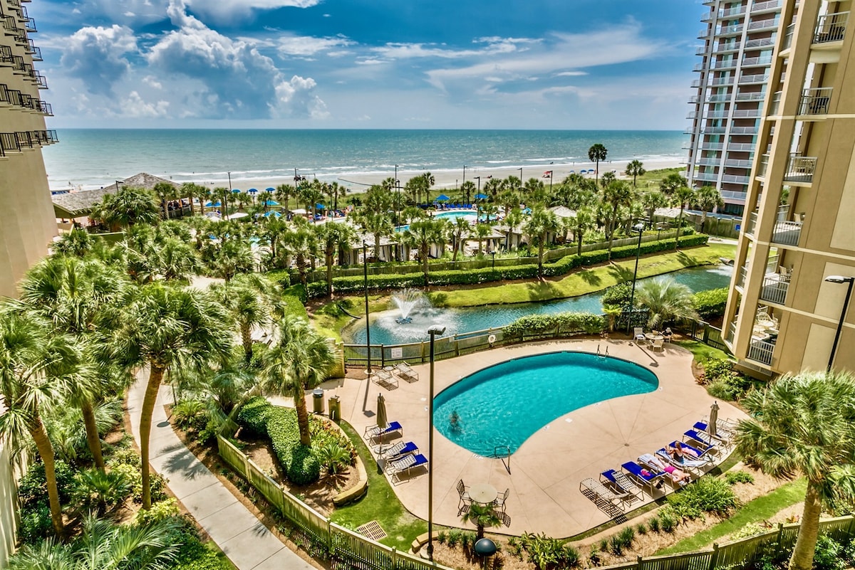 An expansive view showcases a serene outdoor pool surrounded by landscaped greenery and palm trees. The tranquil ocean is visible in the background, and sun loungers are positioned neatly around the pool area, inviting relaxation. The scene reflects a calm coastal atmosphere.