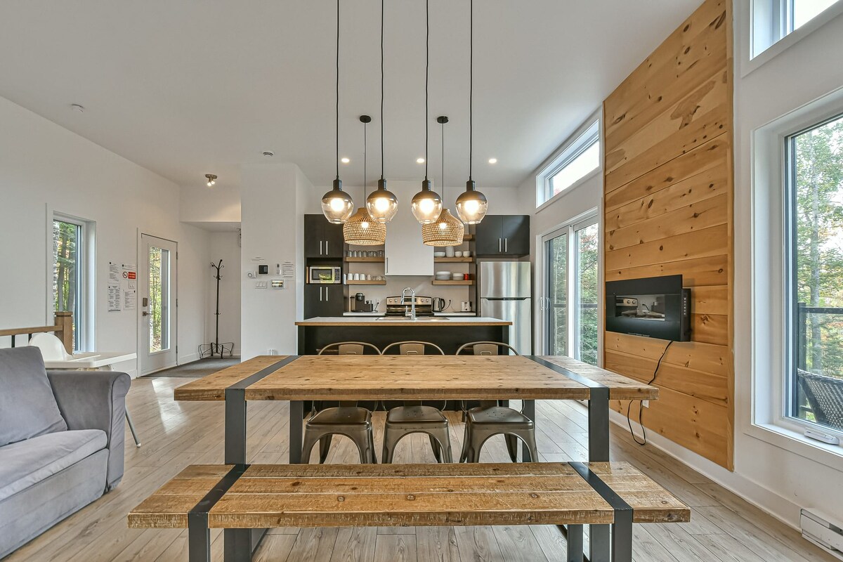 An open-concept kitchen and dining area is featured, with a wooden dining table accompanied by metal chairs. Modern appliances, including a refrigerator and oven, are visible in the kitchen. Large windows allow natural light to fill the space while offering views of the surrounding woodland.