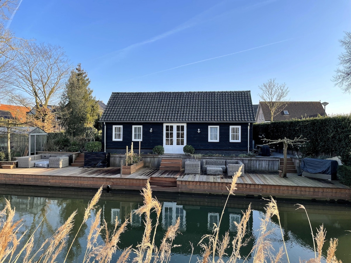 A charming black wooden chalet is positioned beside a tranquil waterway, with a well-designed wooden terrace extending alongside. Several lounge areas are present, offering comfortable seating. Surrounding greenery is visible, creating a serene escape complemented by clear blue skies.