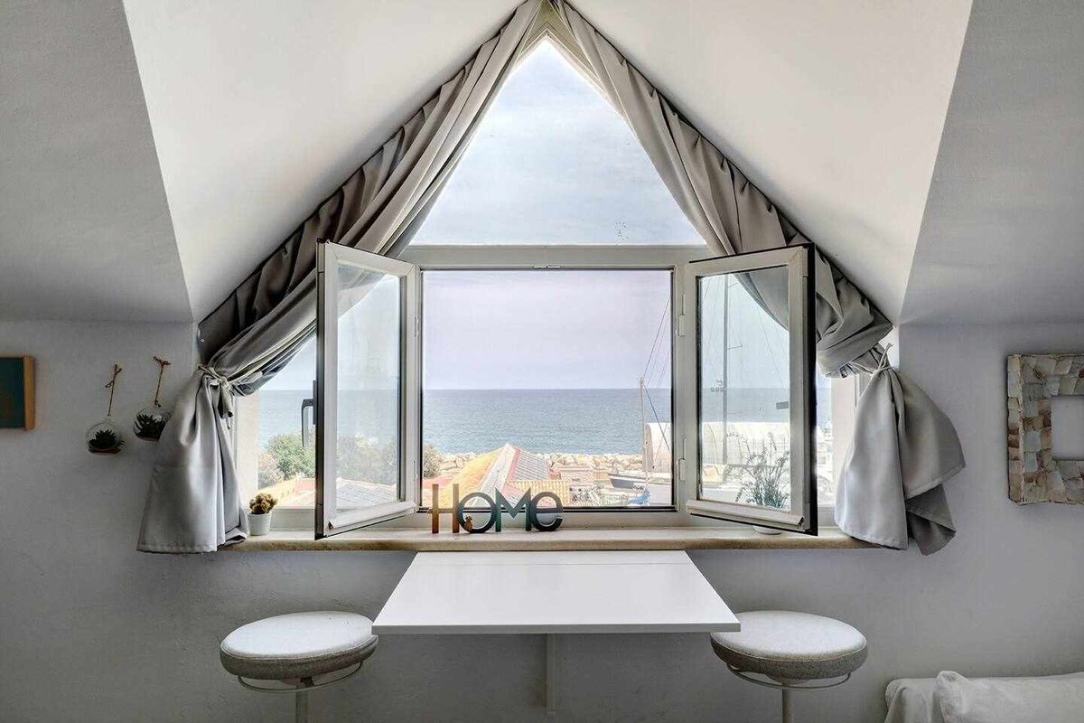 A triangular window reveals a broad view of the sea, framed by elegant grey drapes. A small table with two bar stools sits beneath the window, creating a quiet nook illuminated by natural light.