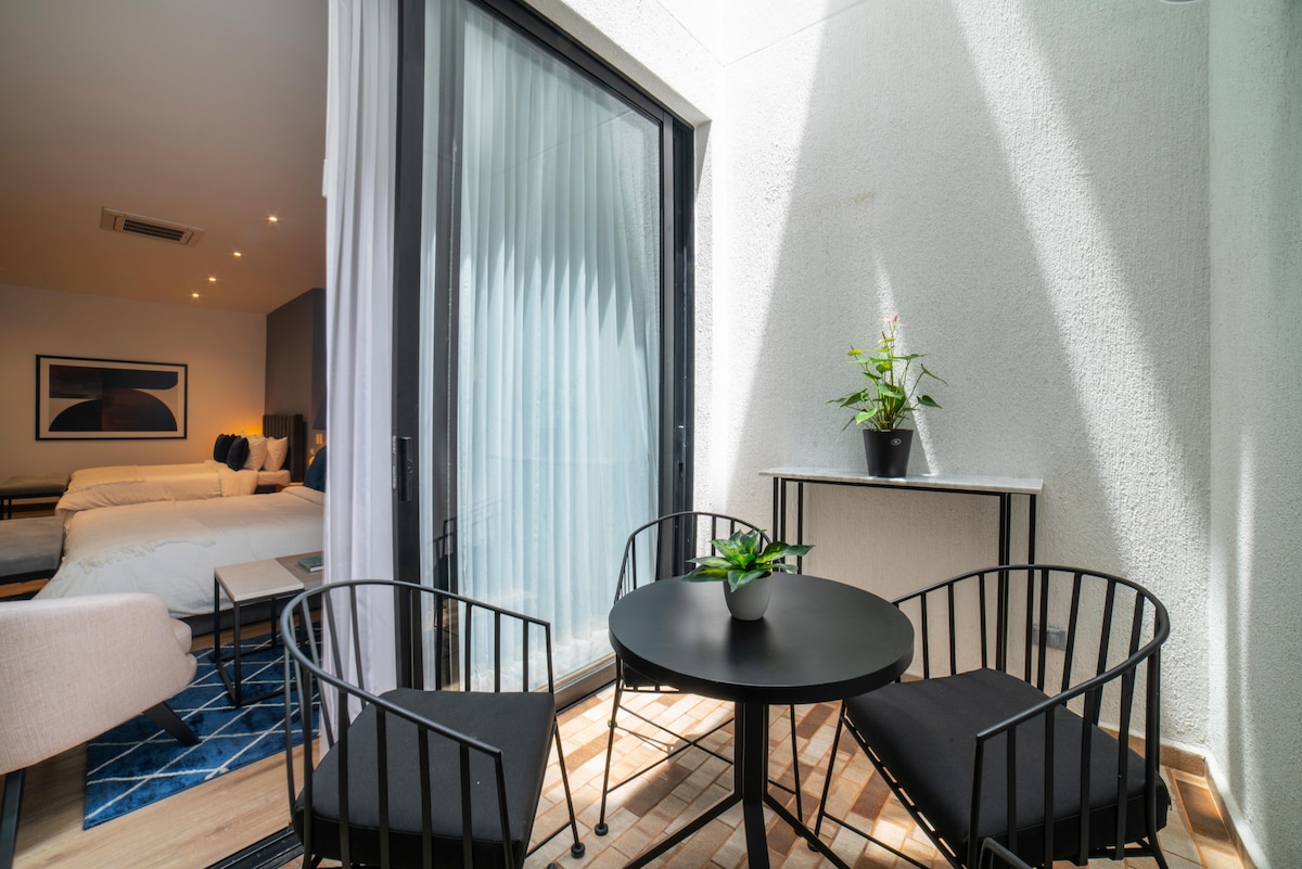 A terrace features a round black table with two matching chairs. A small plant adds a touch of greenery. Natural light filters through large glass doors leading to a bedroom, enhancing the space's openness.