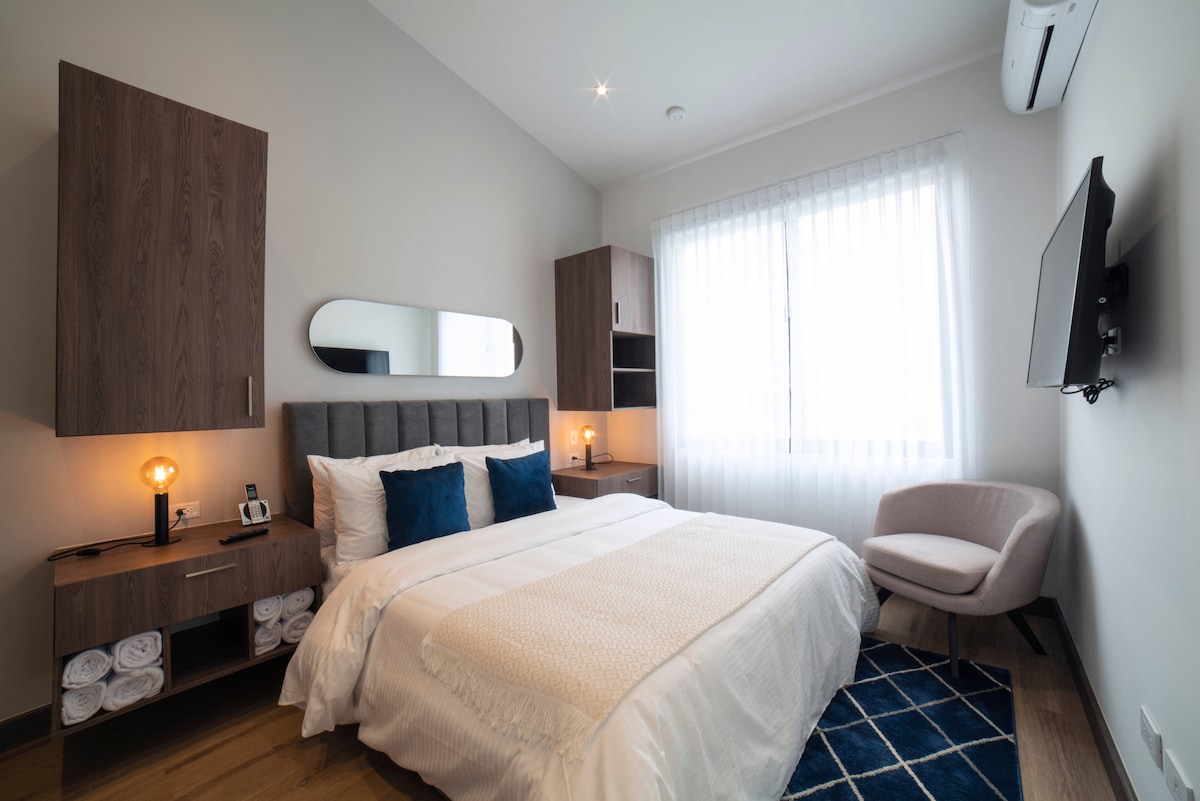 A bright bedroom features a spacious bed adorned with a light-colored blanket and blue accent pillows. Light wood cabinets and a soft rug enhance the space. A comfortable chair is positioned near a window covered with sheer curtains, and a television is mounted on the wall.