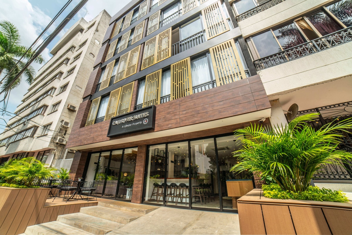 A modern apartment building is showcased with large glass windows and a welcoming entrance. Wooden accents and vibrant greenery enhance the facade. An outdoor seating area is visible, providing space for guests to relax and enjoy the surroundings.