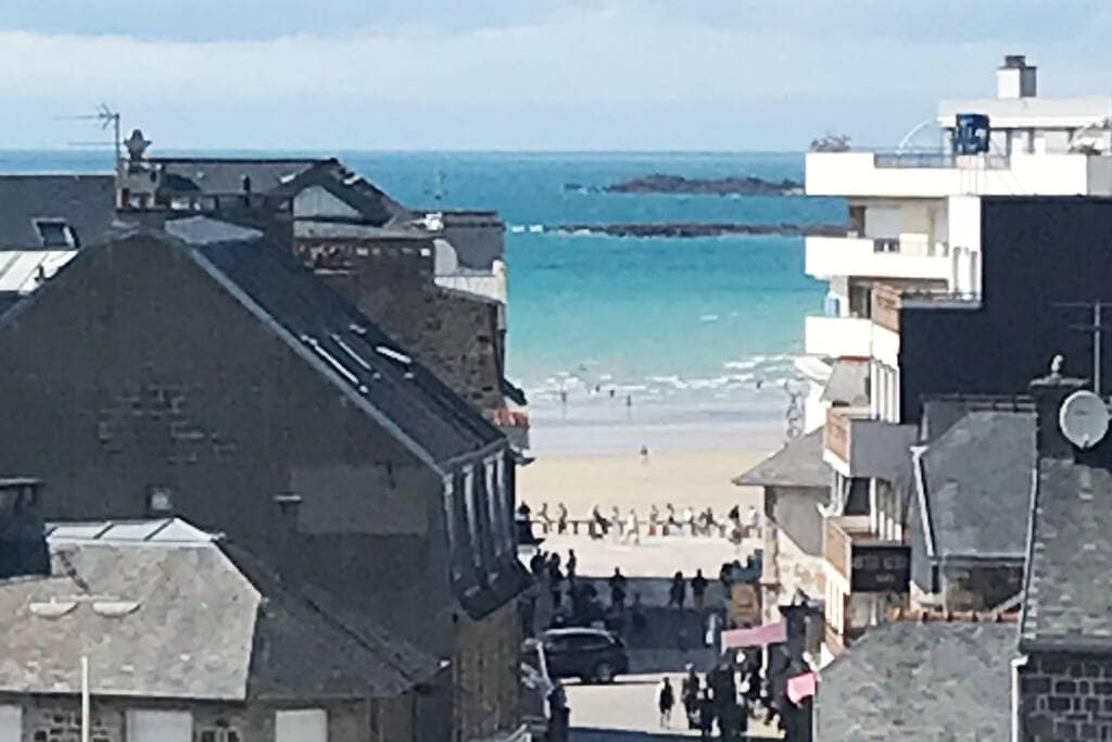 A view of the coastline reveals sand and waves with distant beachgoers enjoying the shore. The scene is framed by charming buildings with slate roofs, enhancing the coastal atmosphere. Light blue ocean contrasts with the sandy beach under a clear sky.