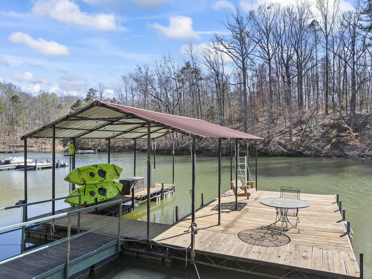 A covered dock presents two green kayaks ready for use. The wooden deck hosts a table with chairs and a bench, surrounded by calm water and tree-lined banks, enhancing the serene lakefront atmosphere.