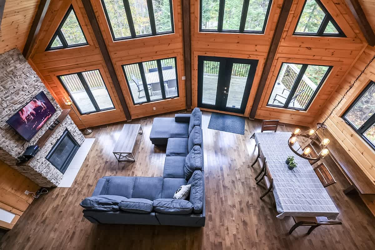 The living area features a comfortable sectional sofa facing a stone fireplace and a wall-mounted television. Large windows allow natural light to flood the space, while a dining table and chairs provide seating for meals. Sliding doors lead to a private outdoor deck.
