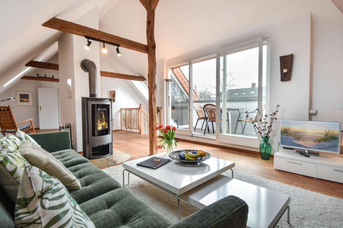 A bright living area features a comfortable green sofa paired with a sleek coffee table. A stylish wood-burning stove adds warmth, while large glass doors invite natural light and provide access to a balcony. A serene landscape painting enhances the space, with fresh flowers completed on the table.