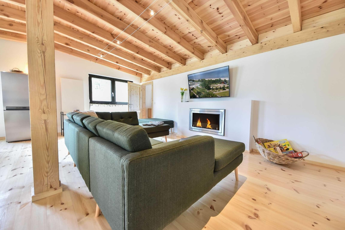 A spacious living area features a comfortable dark green couch positioned near a modern bio-ethanol fireplace. Natural wooden beams frame the ceiling, adding warmth to the space. A flat-screen TV is mounted on the wall, and sunlight pours through large windows.