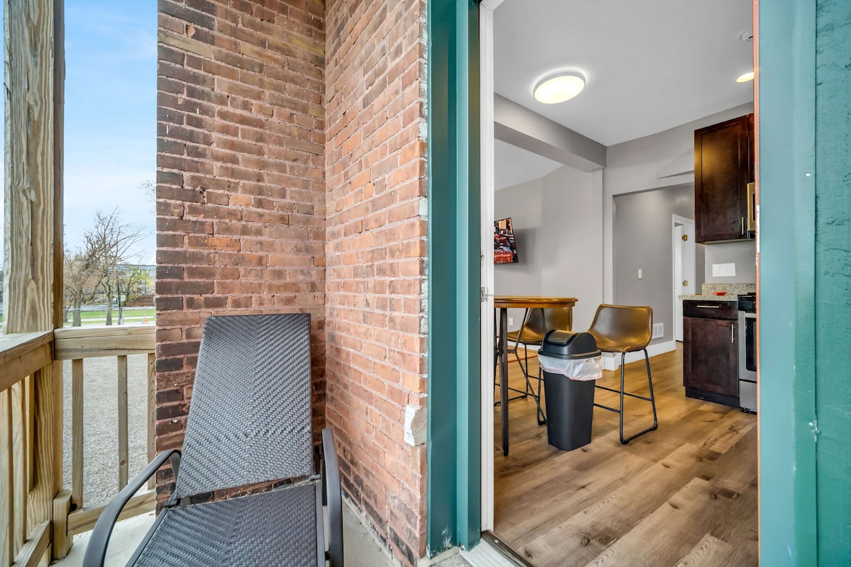 An entrance is framed by exposed brick walls, leading to a bright dining area with modern furnishings. A comfortable chair sits on a small outdoor balcony, with views of the surrounding area. Natural light creates a welcoming atmosphere in the space.