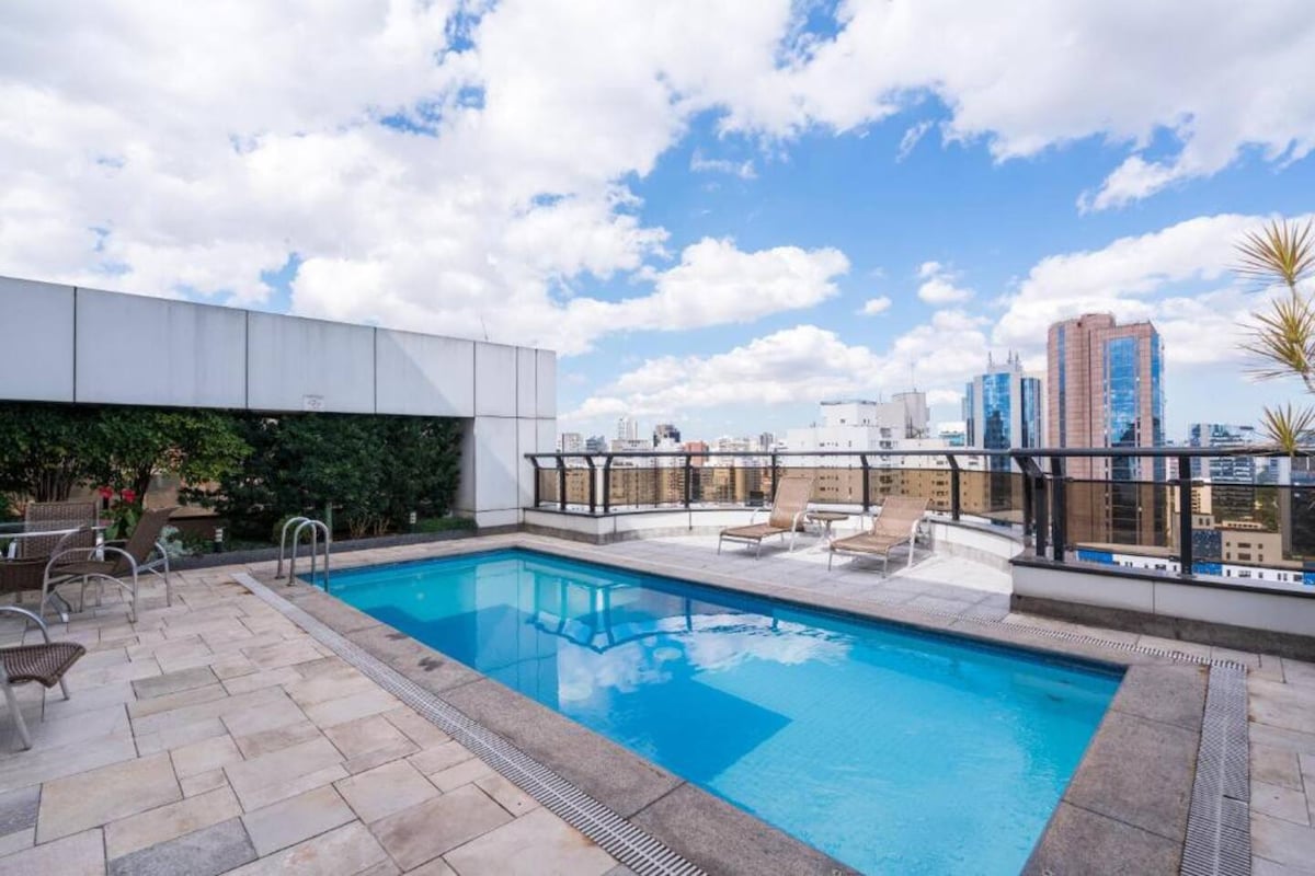 A rooftop pool area is visible, featuring a rectangular swimming pool surrounded by a spacious deck. Comfortable lounge chairs are arranged alongside, with a backdrop of city buildings and a partly cloudy sky.