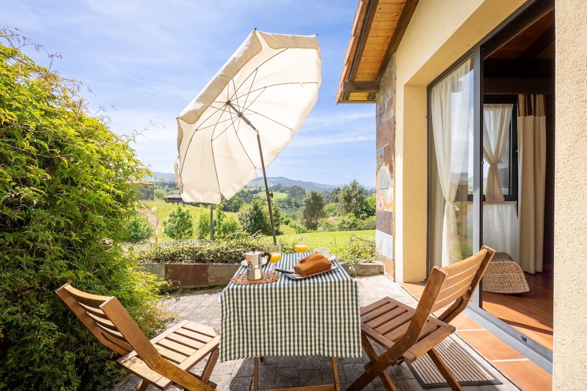 A private terrace features a checkered tablecloth set with a coffee cup and a loaf of bread. Two wooden chairs are positioned around the table, and a large umbrella provides shade. Lush greenery and rolling hills are visible in the background.