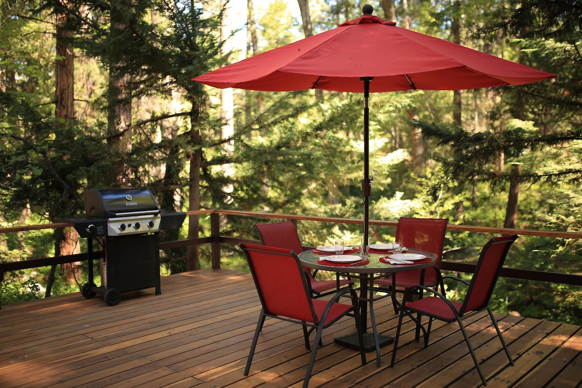 An outdoor deck features a round table with four red chairs and a large red umbrella providing shade. A propane grill stands nearby, surrounded by tall trees that create a serene and secluded atmosphere.