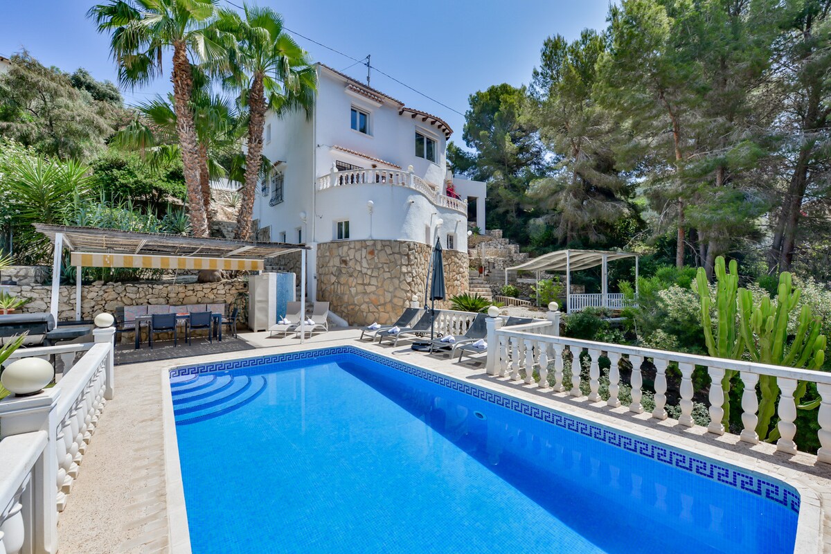 A spacious outdoor area is showcased, featuring a private swimming pool surrounded by palm trees and lounge chairs. Various terraces and shaded seating areas can be seen, along with stone walls that enhance the natural surroundings of the villa.