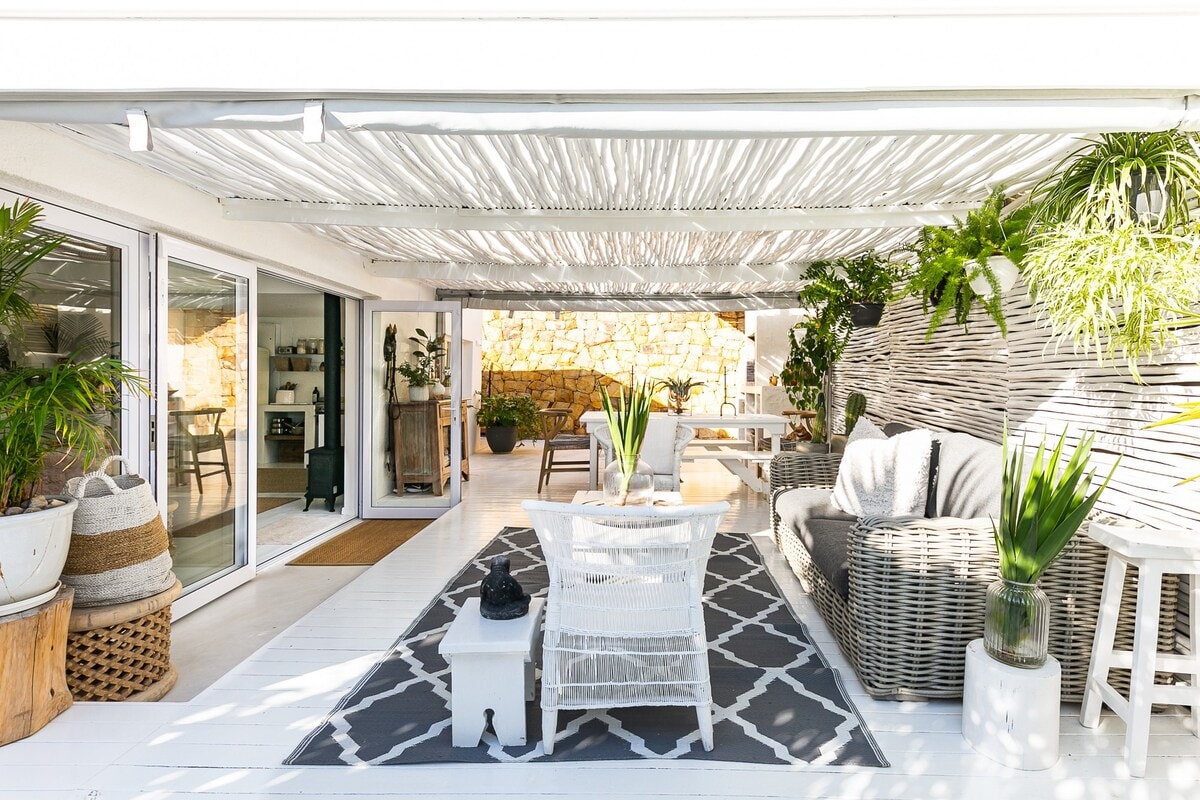 A spacious patio is shaded by a textured overhead covering, with potted plants adding natural elements. Comfortable seating includes a woven sofa and chairs, surrounded by a neutral-colored rug. Light filters in through large glass doors, enhancing the airy ambiance.
