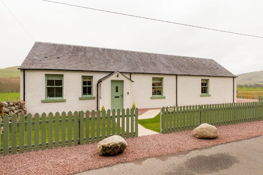 The exterior of Balbeg Cottage is presented, showcasing a one-story building with a neutral facade and green window frames. A gravel pathway leads to the entrance, bordered by a low picket fence and two large stones at the front.