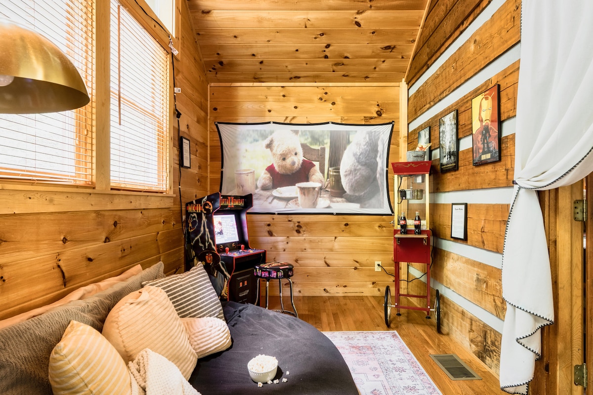 A cozy area is presented with wooden walls and large windows allowing natural light. A big screen displays an animated scene, and a vintage popcorn cart sits nearby. A retro arcade game station is positioned to one side, with a plush sleeping space adorned with decorative pillows.