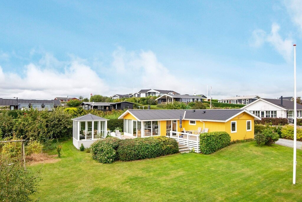A holiday home is depicted in a vibrant yellow hue, set within a lush green garden. Surrounding structures are visible in the background, along with trees and well-maintained hedges. A covered terrace extends from the house, offering outdoor seating options and inviting views of the landscape.