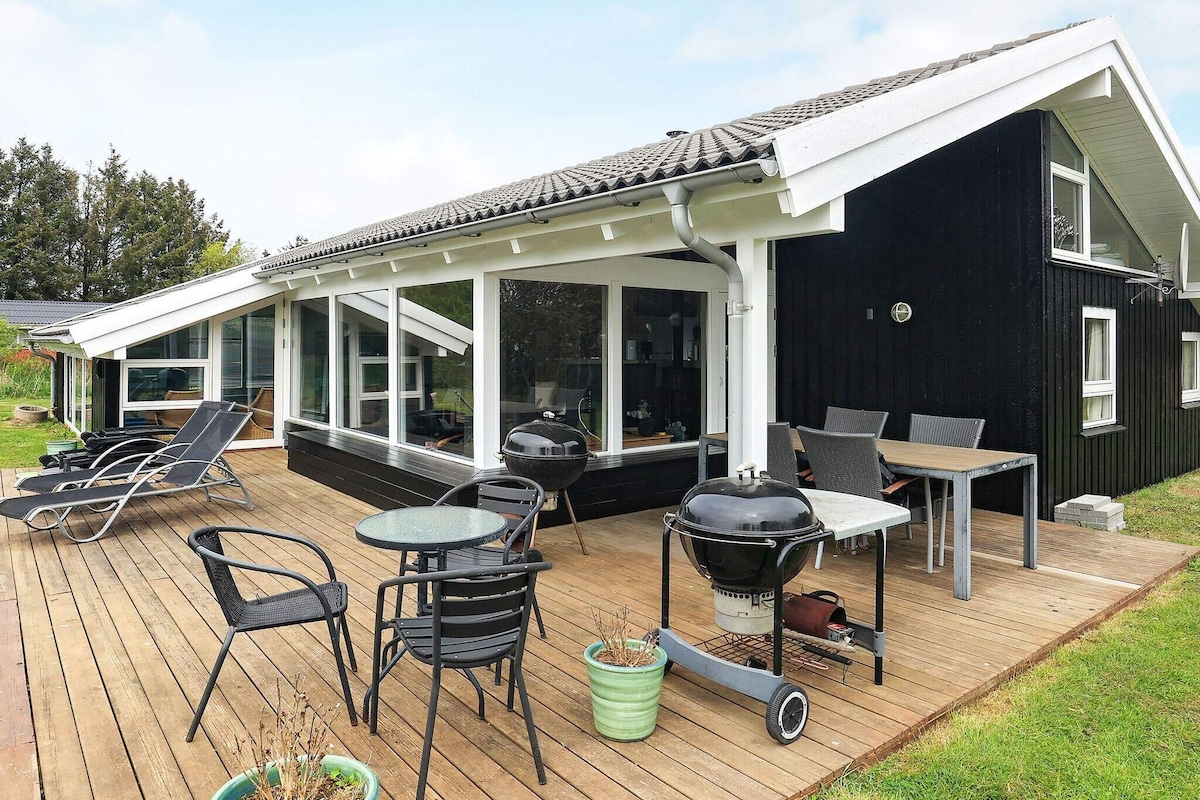 An outdoor terrace features a mix of seating options, including lounge chairs and dining furniture, inviting relaxation. A barbecue grill is placed conveniently nearby, with planters seen in the foreground. Large windows provide a view of the natural surroundings.