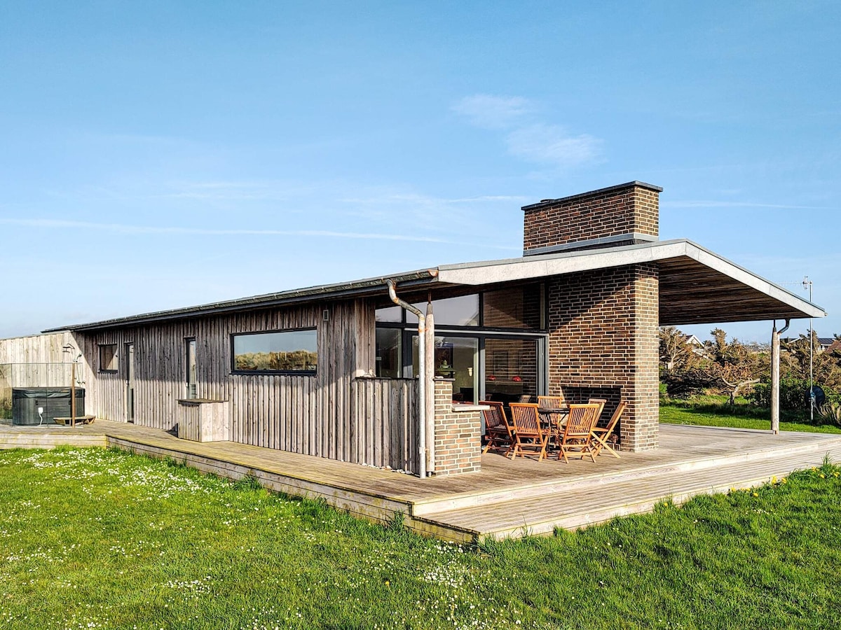 A modern wooden holiday home features a spacious terrace with seating arrangements. Large windows provide views of the surrounding landscape, while a chimney adds character. The exterior is complemented by grassy areas and a serene atmosphere, ideal for enjoying outdoor activities.
