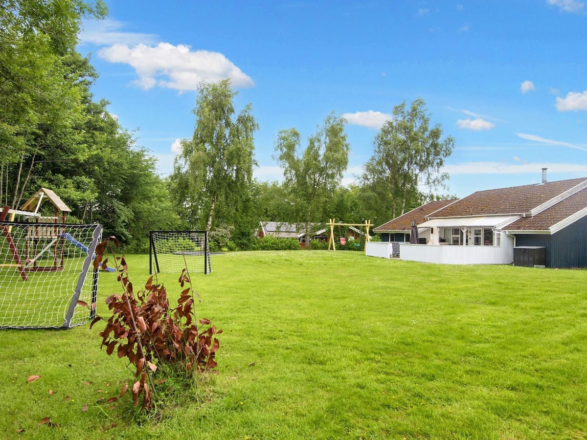 A spacious lawn area is depicted, featuring several children's play structures and two football goals. The well-maintained grass is surrounded by trees, providing a sense of privacy. The house is visible in the background, complemented by a clear blue sky above.