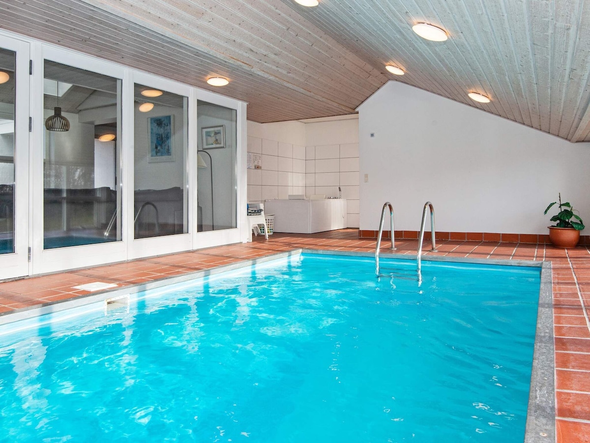 An indoor swimming pool is visible, featuring clear blue water and steps leading in. Natural light from large glass doors illuminates the space, creating a bright environment. Adjacent to the pool area, a jacuzzi is partially seen, providing an opportunity for relaxation.