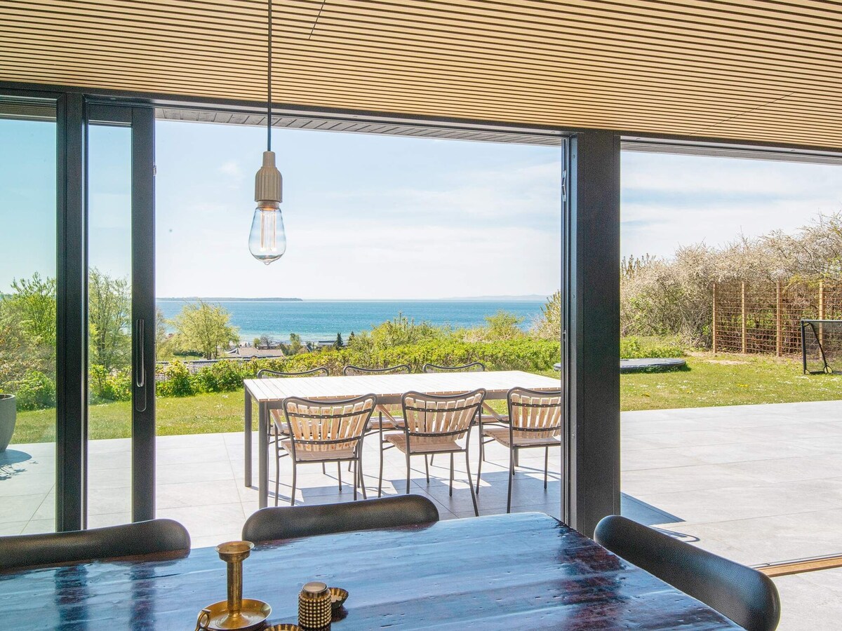 Large glass doors provide a seamless view of the outdoor terrace and the water beyond. A dining table with several chairs is positioned on the terrace, surrounded by greenery. The space is illuminated by soft natural light, enhancing the airy atmosphere.
