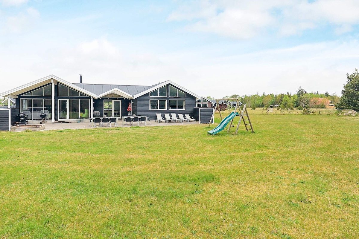 A spacious holiday home is presented with a broad lawn surrounding it. Sun loungers are arranged on the patio, and a children's play area features a slide and swings. The minimalist facade includes large windows that invite natural light into the interiors.