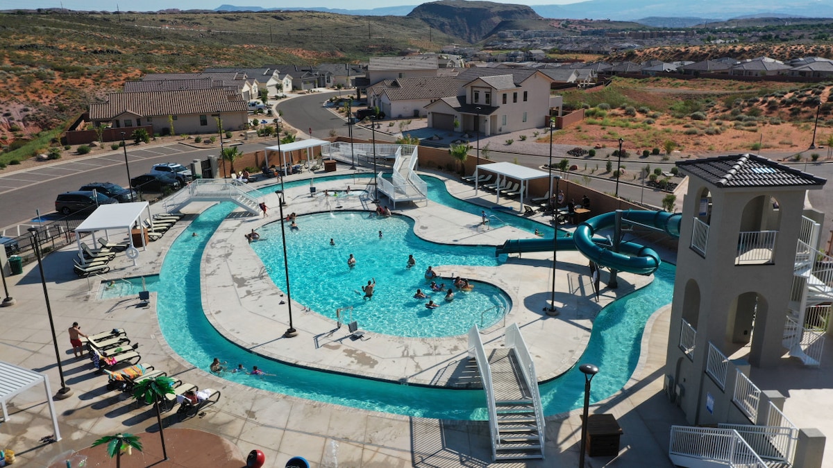 An aerial view showcases a large pool area featuring a winding water slide and several lounge chairs arranged under shaded pergolas. Families and individuals enjoy the water, with surrounding homes and desert landscape visible in the background.