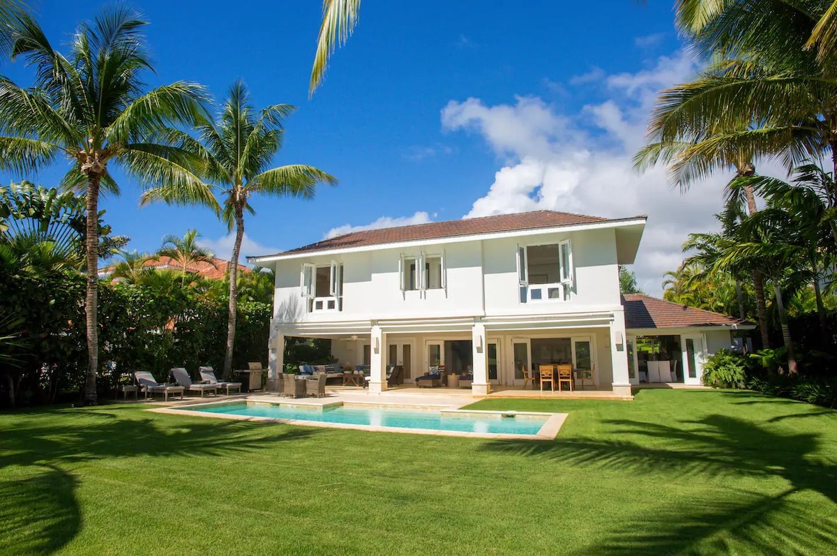 A two-story villa is surrounded by lush palm trees and well-maintained greenery. The exterior features large windows and a patio area that overlooks a private pool, with seating arrangements nearby. The blue sky adds a vibrant backdrop to the serene setting.
