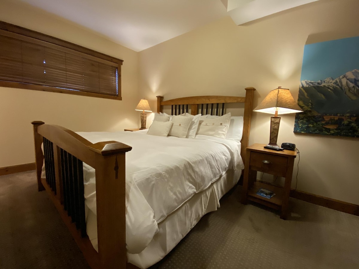 A cozy bedroom features a spacious bed with crisp white linens and plush pillows. Two bedside lamps provide soft illumination. A wooden bed frame complements the rustic decor, while a window with wooden blinds allows for privacy and control of natural light.