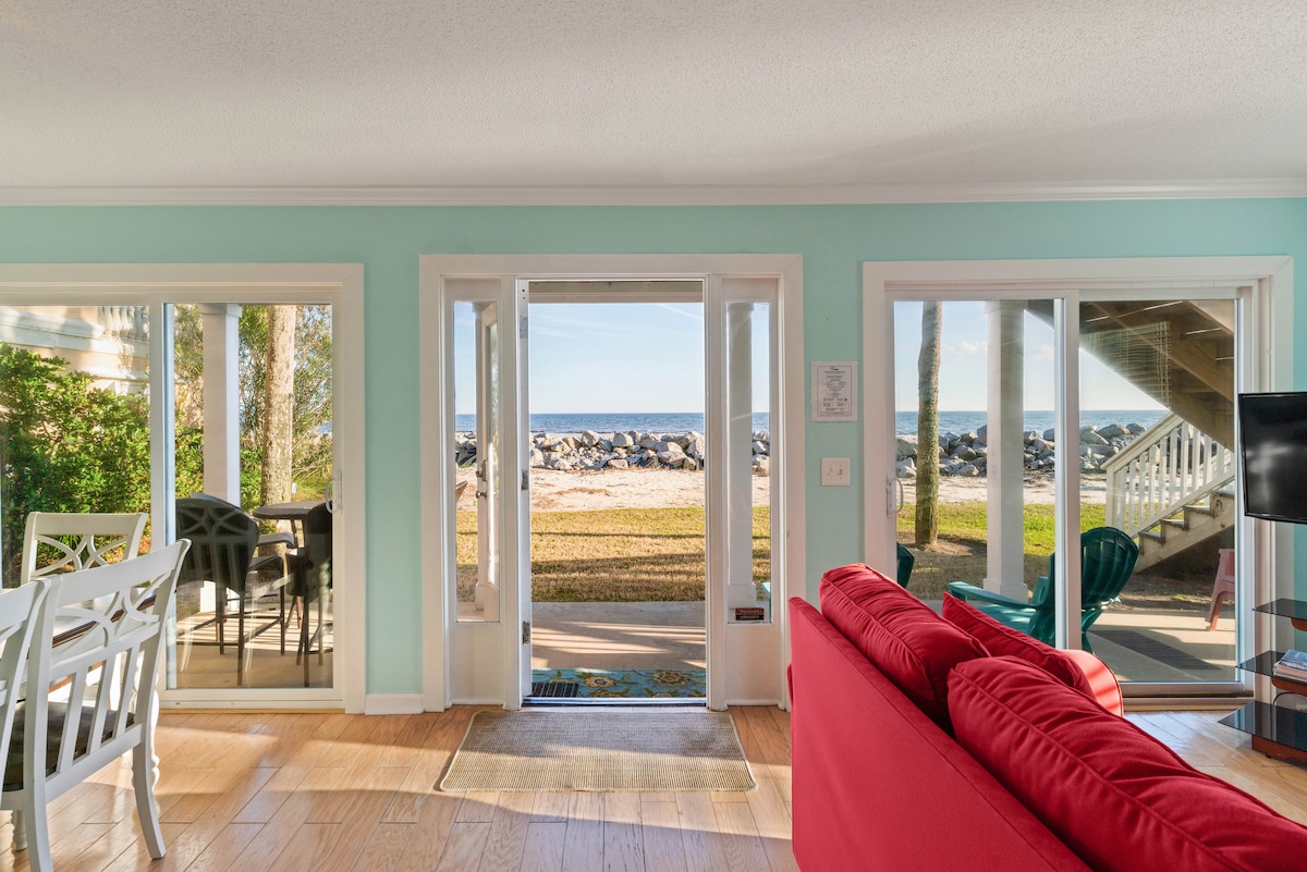 A spacious living area is visible through open glass doors, allowing unobstructed views of the Atlantic Ocean and beach. A bright red sofa adds a pop of color to the room, complemented by light wood flooring and aqua walls. The outdoor space features a lawn and rocky beachfront.