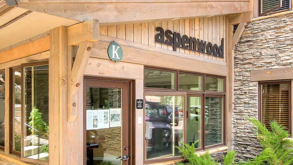 The exterior of the Aspenwood complex is showcased, featuring warm wooden accents and stone detailing. Large windows allow natural light to flood in, while a welcoming sign identifies the entrance. Lush greenery surrounds the entryway, adding to the inviting atmosphere.