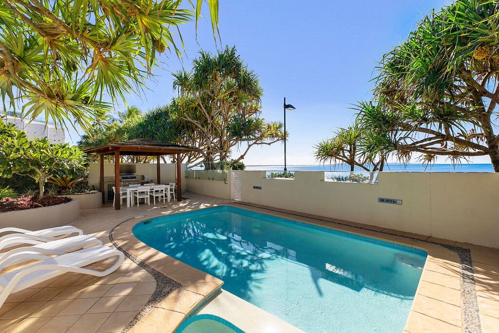 An outdoor swimming pool is surrounded by tropical greenery, offering a relaxing atmosphere. A shaded seating area with a dining table is visible nearby. The ocean can be seen in the background, enhancing the tranquil ambiance of the space.