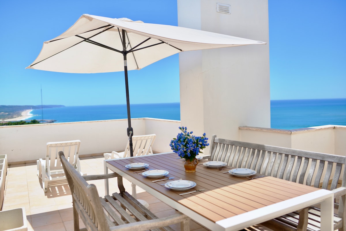 An outdoor terrace features a wooden dining table set for meal times, accompanied by eight plates and a vase with blue flowers. A large umbrella provides shade, while two sun-loungers are positioned nearby, offering views of the coastline and the ocean.
