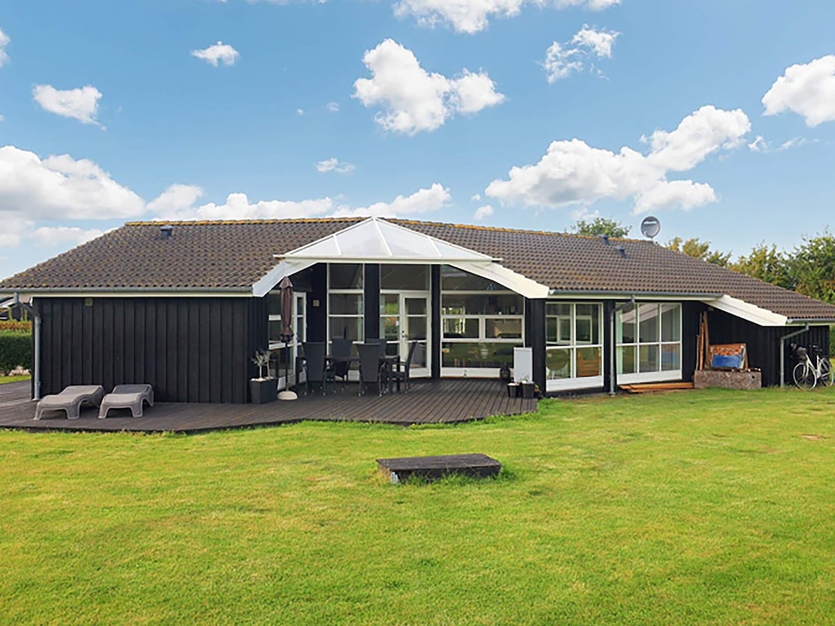 A modern holiday home is shown, featuring a large overhanging roof and expansive windows. The building is surrounded by a well-kept grassy area, with outdoor seating visible on a spacious deck. Nearby, bicycles can be seen, adding to the leisure atmosphere.