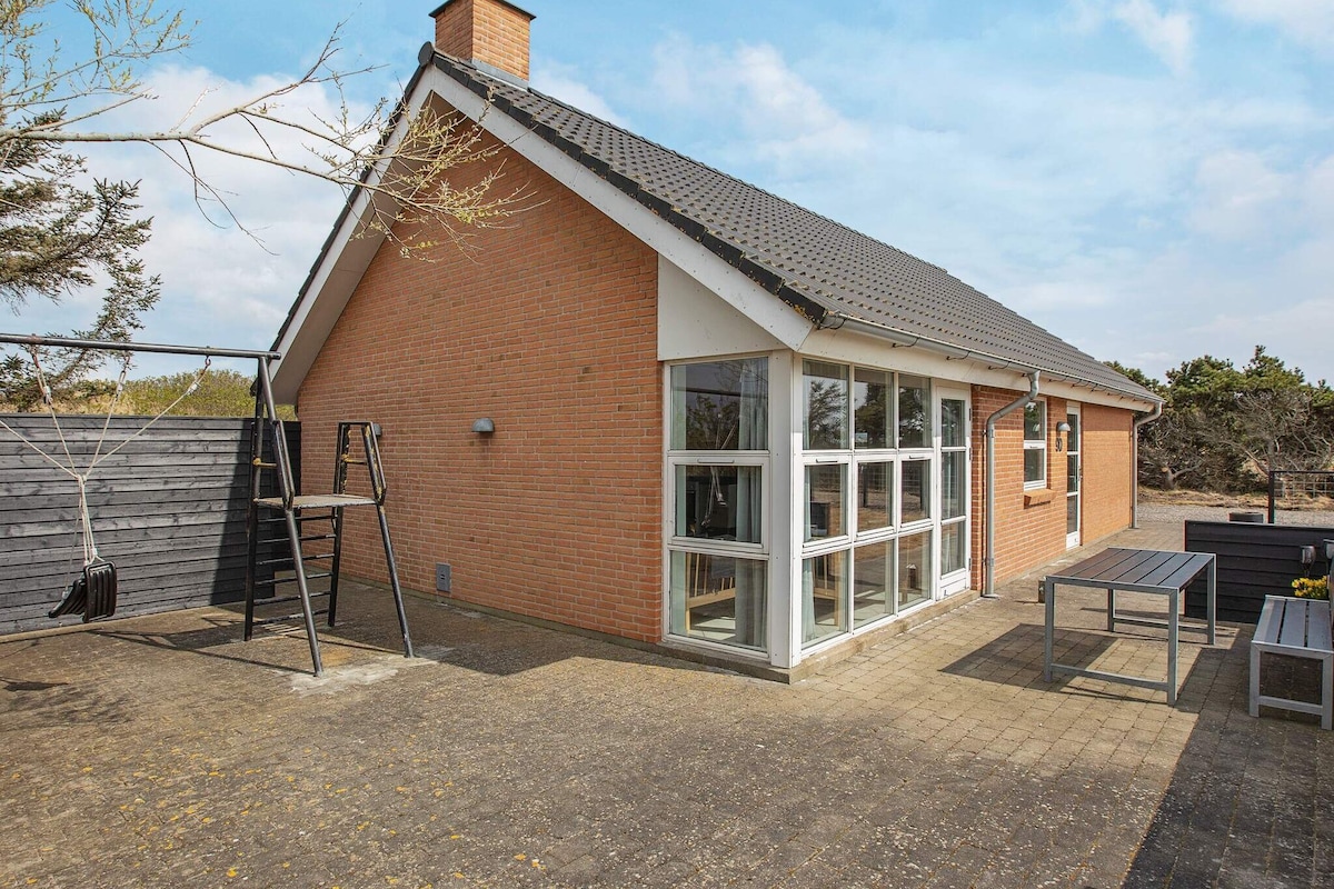 The exterior of the holiday home showcases a modern brick façade with large windows allowing natural light to illuminate the interior. A spacious, paved patio area features outdoor furniture and a swing set, surrounded by a mix of greenery and open space.