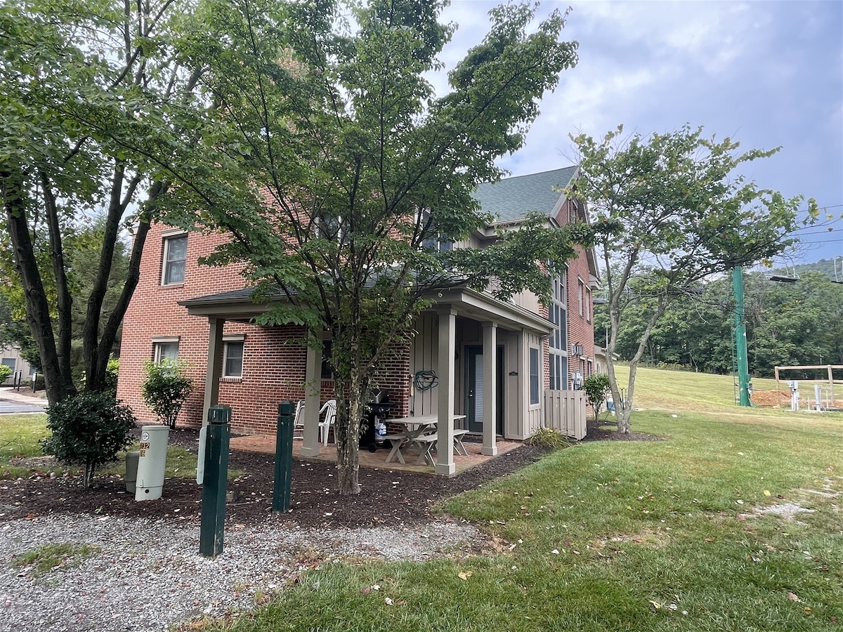 The exterior of the townhouse is highlighted, featuring a brick façade surrounded by trees and greenery. A covered porch area offers seating, while the landscaped yard includes gravel pathways and grassy areas. The structure stands two stories tall with large windows visible.
