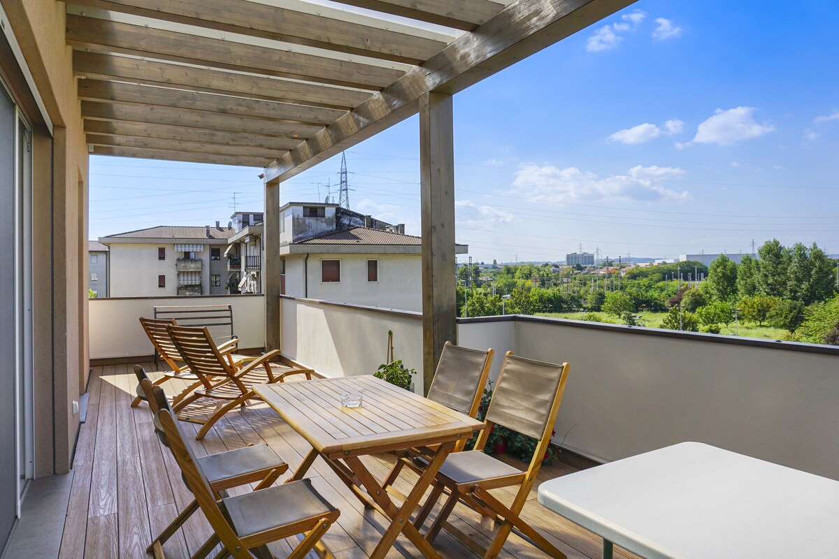 A spacious terrace features a wooden pergola overhead, with a large dining table surrounded by six wooden chairs. The area overlooks greenery and distant buildings under a clear blue sky, providing an inviting space for outdoor relaxation or meals.