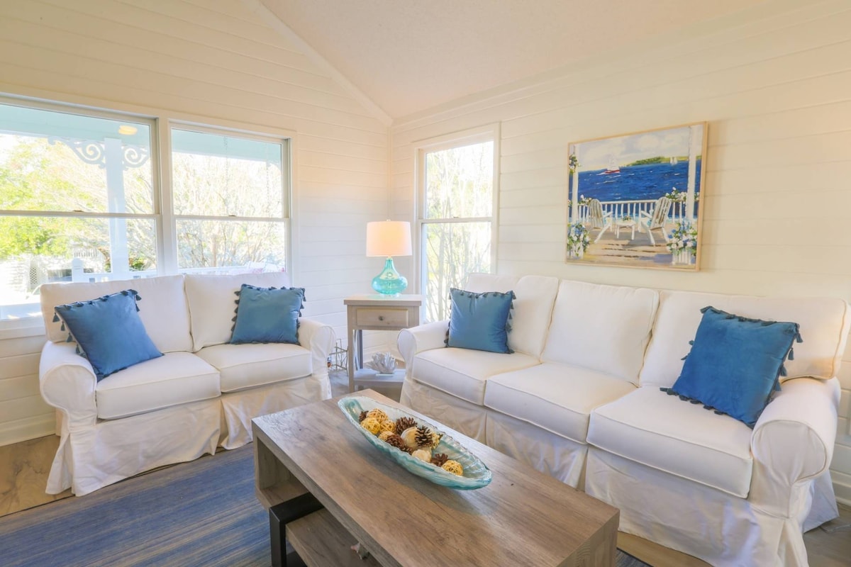 A comfortable living room features two white sofas adorned with blue accent pillows. A medium wooden coffee table, holding a decorative bowl with seashells, sits in front. Large windows allow natural light, highlighting the shiplap walls and creating a bright atmosphere.