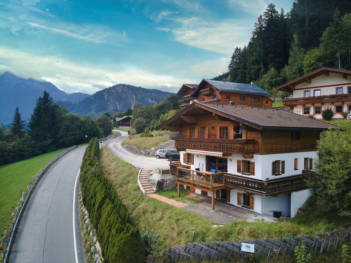 A charming chalet is nestled among lush greenery, overlooking a winding road and mountains. The structural elements include wooden balconies and a landscaped garden area. The surrounding natural scenery features tall trees and distant peaks under a lightly clouded sky.