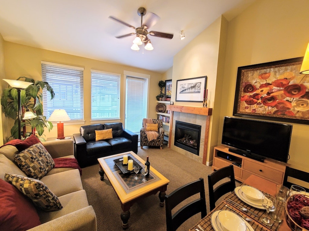 The living area features a comfortable sofa and armchair arranged around a central coffee table. A flat-screen TV and a cozy fireplace enhance the inviting atmosphere. Natural light filters through the large windows adorned with blinds, providing a well-lit space for relaxation.